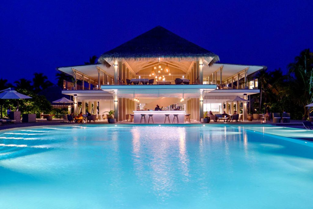 Bar im Baglioni Resort Maldives mit Sitzgelegenheiten unter Sonnenschirmen und beleuchtetem Pool in der Dämmerung