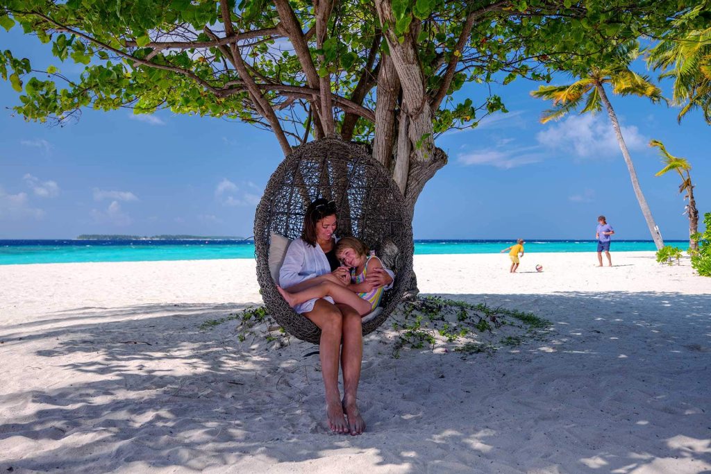 Zwei Personen sitzen in geflochtenem Sitzkorb unter Baum am weißen Sandstrand mit Meer und blauem Himmel im Hintergrund