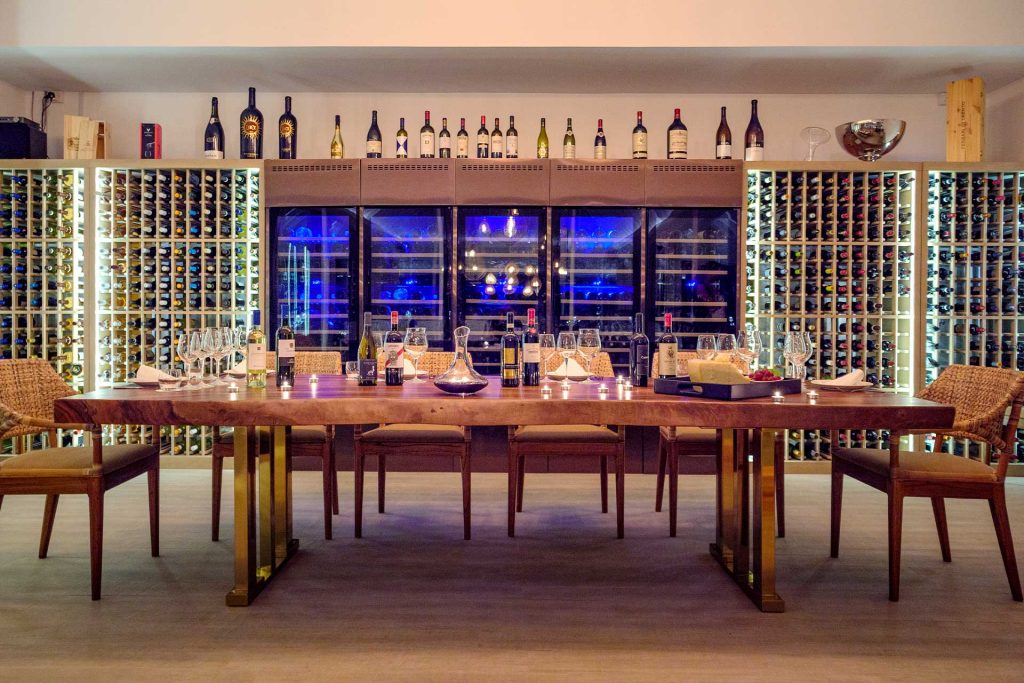 Weinkeller im Baglioni Resort Maldives mit Holztisch, Rattanstühlen, Weinflaschen, Gläsern und beleuchtetem Weinkühlschrank