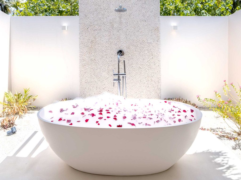 Freistehende Badewanne mit schaumigem Wasser und rosa Blüten in einem weißen, von Kieselsteinen bedeckten Außenbereich
