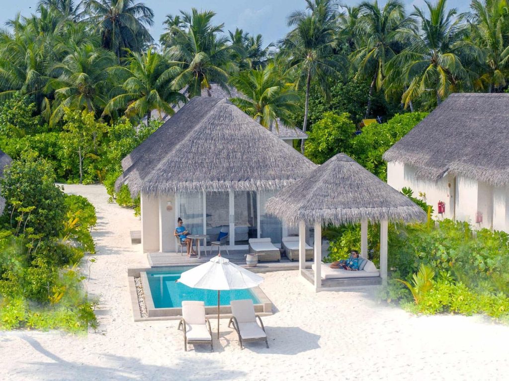 Blick auf Poolbereich mit weißen Liegen und Sonnenschirm neben Bungalows mit Strohdächern im Baglioni Resort Maldives