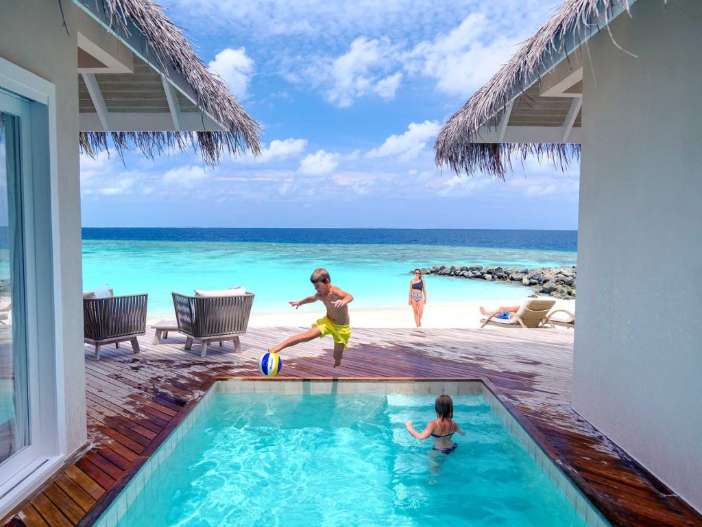 Poolbereich mit Holzdeck und Strohdach am weißen Sandstrand mit Liegestühlen und türkisfarbenem Meer im Hintergrund