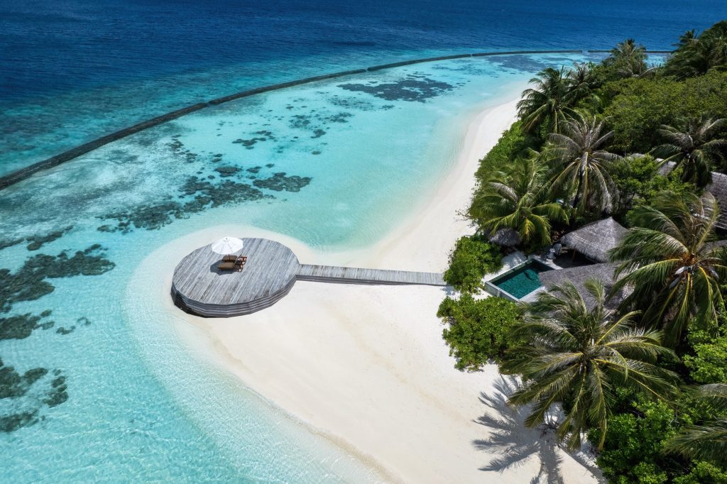 Holzkai mit Liegestühlen und Sonnenschirm am türkisfarbenen Wasser neben weißem Sandstrand und Palmen auf den