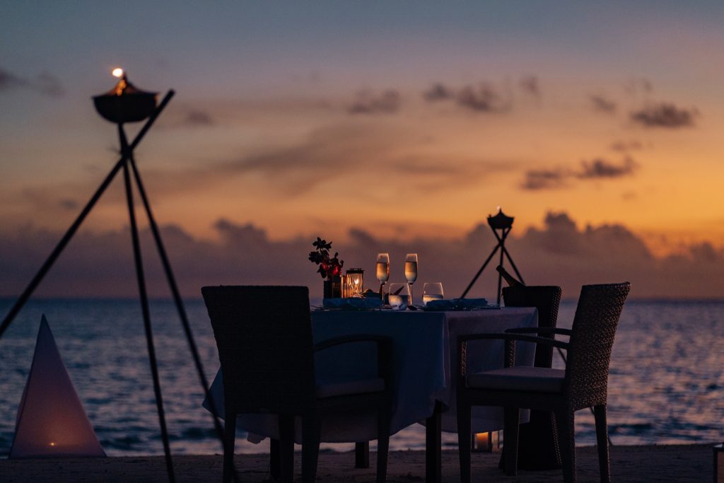 Abendessen am Strand bei Baros Maldives mit Tisch, Rattanstühlen, Kerzen und Sonnenuntergang über dem Meer