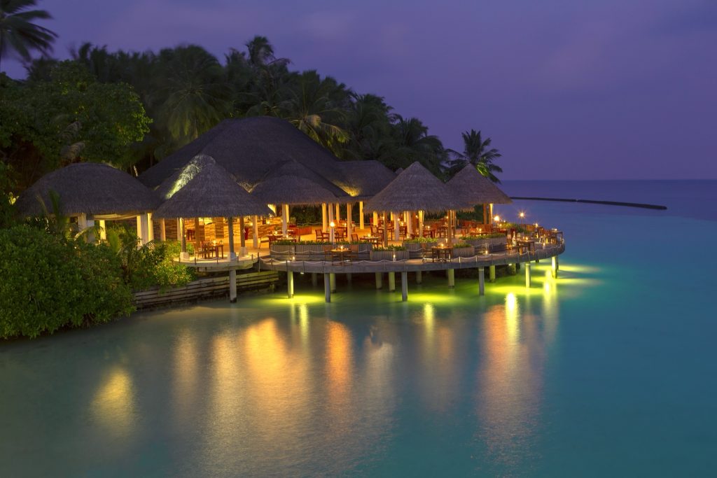 Restaurant Cayenne in Baros Maldives mit strohgedecktem Dach über türkisfarbenem Wasser und Holzsteg bei Abendlicht