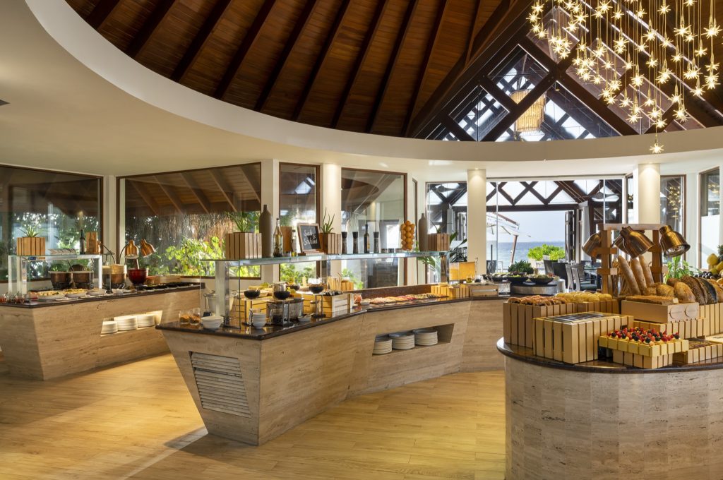 Buffet im Baros Maldives Restaurant Lime mit runden Tischen aus Stein und Holz, Speisen und Meerblick durch große Fenster