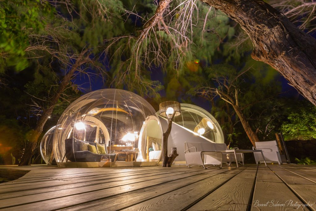 Transparente kuppelförmige Zimmer der Bubble Lodge Île aux Cerfs mit Holzterrasse und Bäumen bei Nacht
