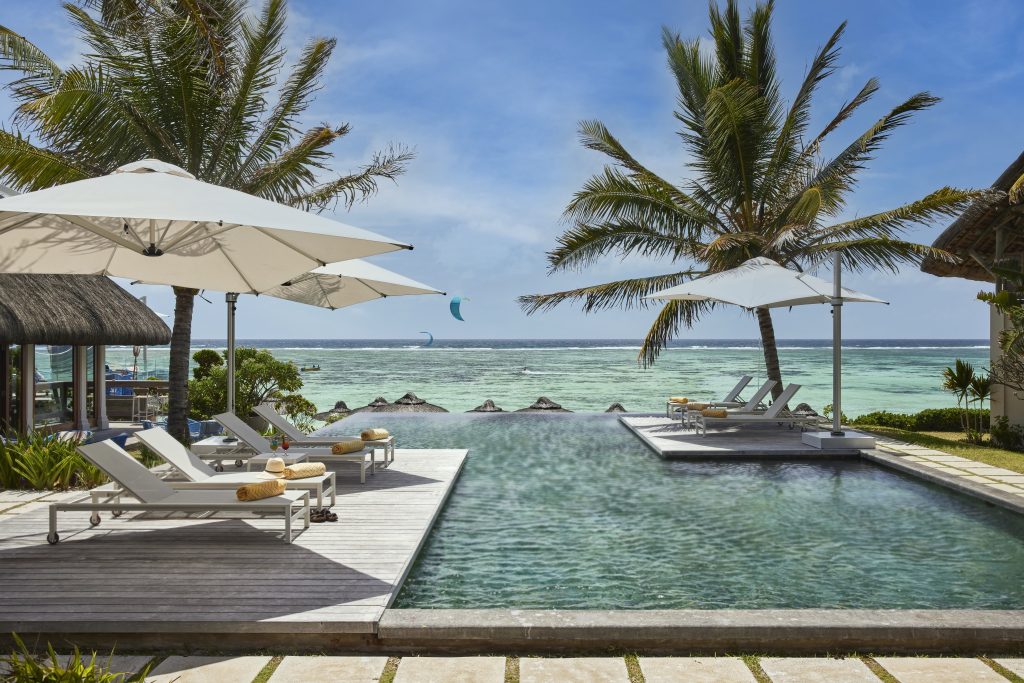 Rechteckiger Pool mit weißen Liegestühlen, gelben Handtüchern und Sonnenschirmen, Palmen und Strand im Hintergrund
