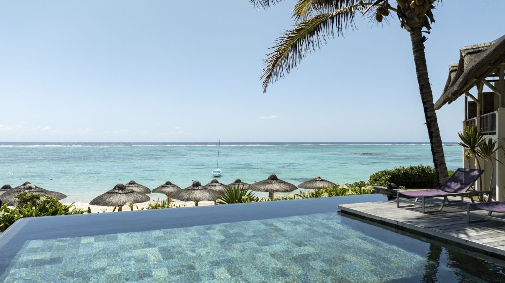 Infinity-Pool am C Mauritius mit Holzumrandung, Palmen, strohgedeckten Sonnenschirmen und Segelboot im Hintergrund
