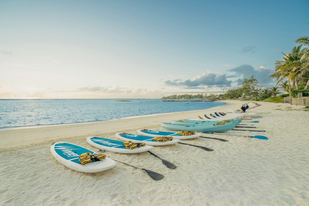 Sandiger Strand mit weißen und blauen Paddleboards und Kajaks, Palmen und Küstenlandschaft im Hintergrund