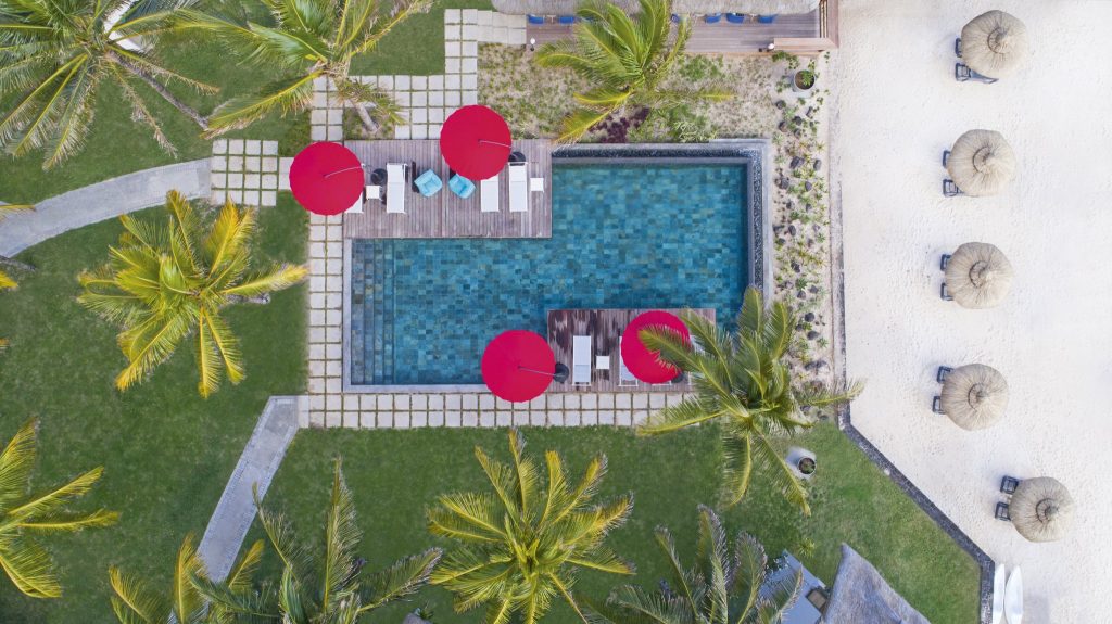 Vogelperspektive auf rechteckigen Pool mit blauen Wasserflächen, Holzdecks, roten Sonnenschirmen und sandigem Bereich.