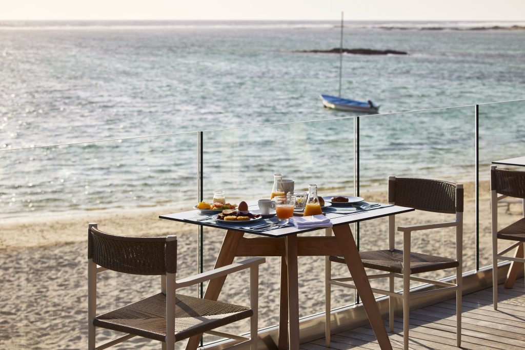 Terrasse mit Holztisch und Glasplatte, Orangensaft, Brötchen, Obst, zwei Holzstühle, Glasgeländer, Meerblick