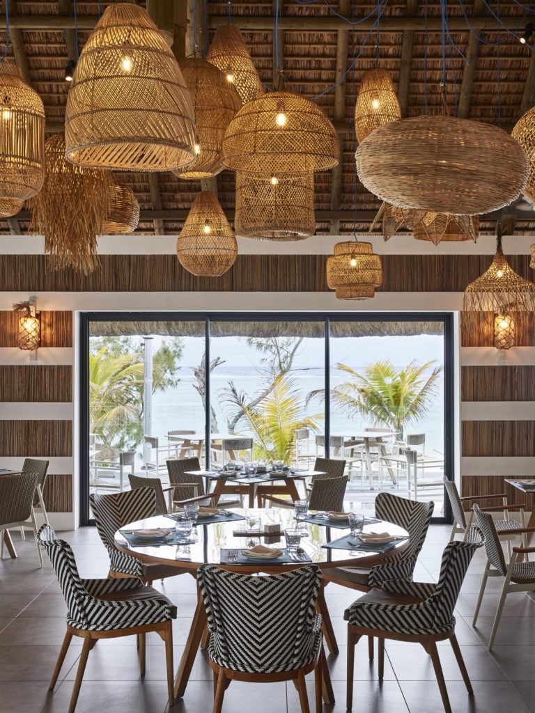 Restaurant im C Mauritius mit strohgedeckter Decke, Rattanlampen, Holz- und Flechtmöbeln, Sicht auf Palmen und Meer