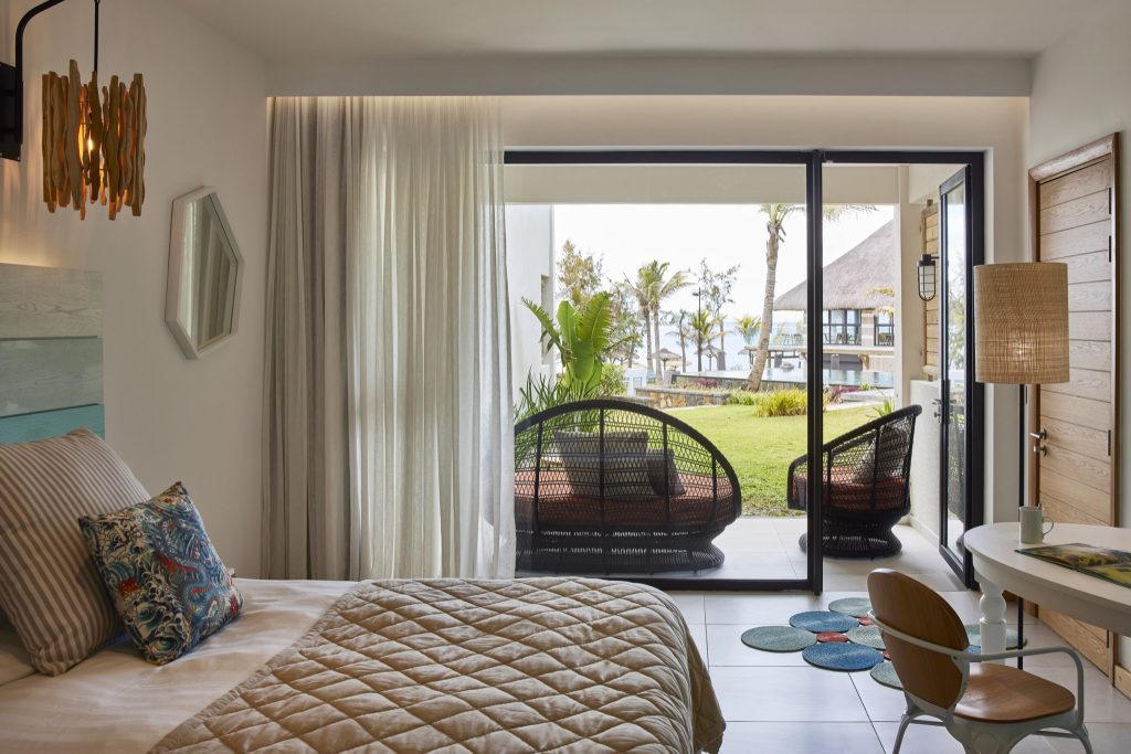 Helles Deluxe Room Hotelzimmer mit Holzmöbeln, gestepptem Bett und Glas-Schiebetür zur Terrasse und Garten mit Palmen