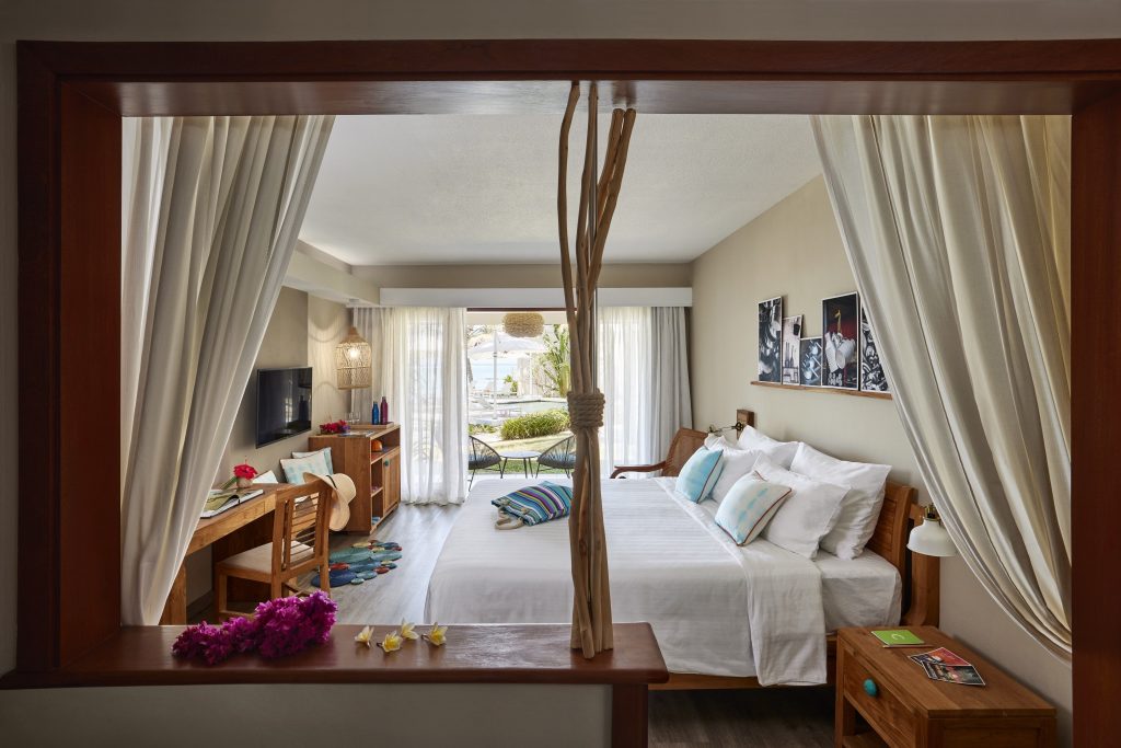 Helles Prestige Room Zimmer mit großem Bett, weißen Bettlaken, blauen Kissen, Holzschreibtisch und Gartenmöbeln draußen