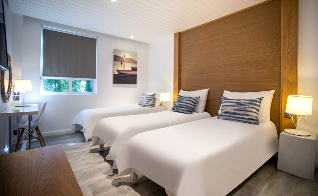 Hotelzimmer mit zwei weißen Einzelbetten, blau-weiß gestreiften Kissen, Holzwand und grauem Rollo in Mauritius