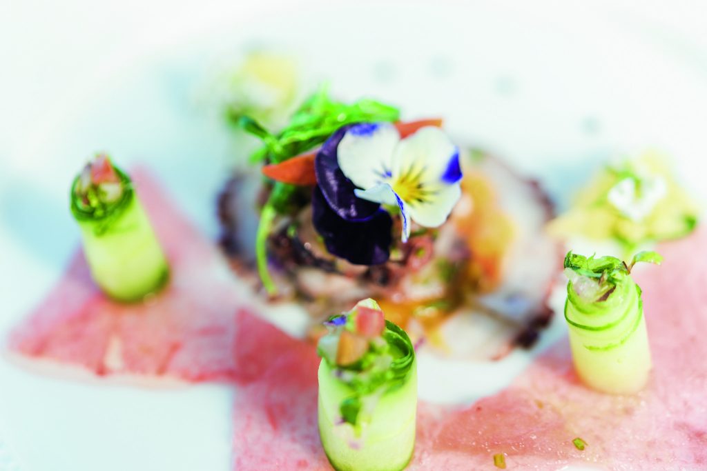 Nahaufnahme eines Tellers mit dünn geschnittenem rosa Fleisch, grünen Gemüserollen und essbaren Blüten im Canonnier.