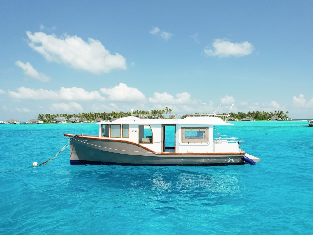 Weißes Boot mit Fenstern und Holzreling auf türkisblauem Wasser vor Insel mit Palmen und Bungalows