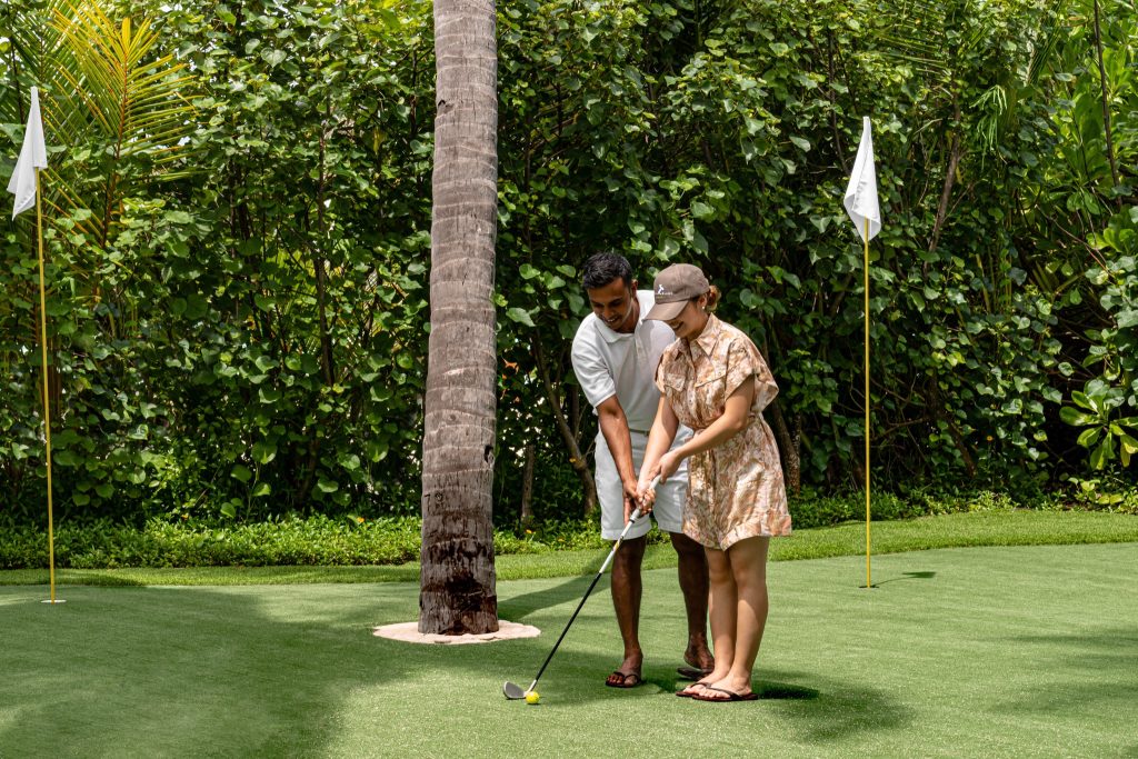Zwei Personen auf grünem Kunstrasen-Golfplatz mit weißen Fahnen und Palmen im Außenbereich des Cheval Blanc Randheli