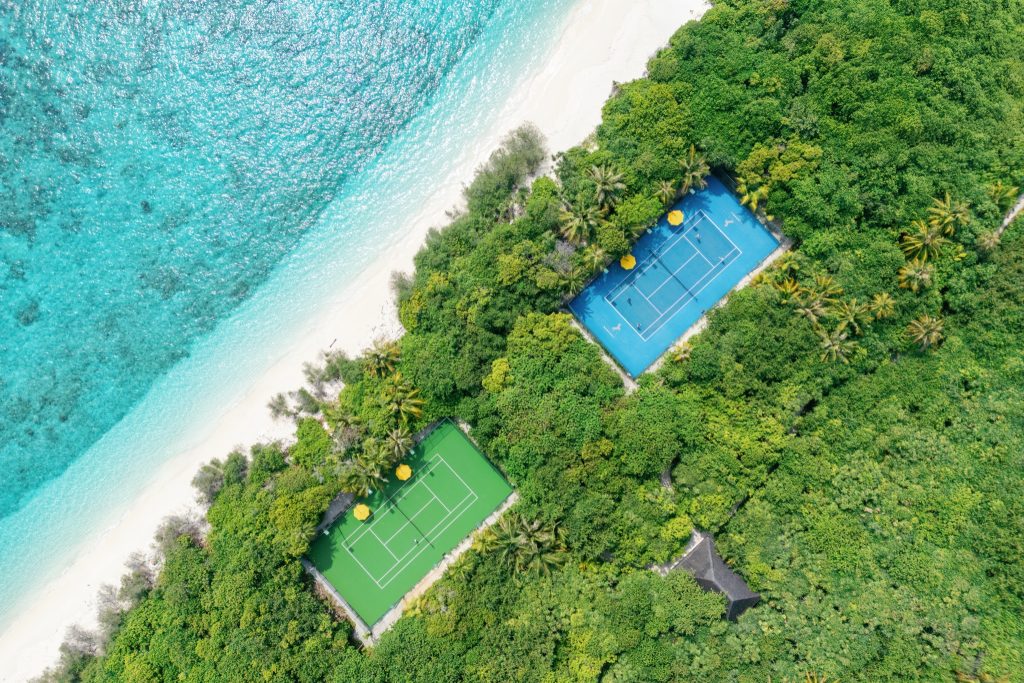 Luftaufnahme von zwei Tennisplätzen mit blauem und grünem Belag, gelben Sonnenschirmen, Strand und Meer auf den
