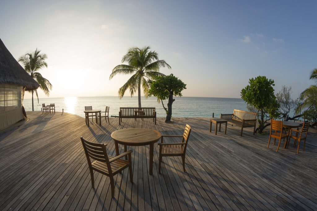 Holzterrasse mit Holztischen und Stühlen, strohgedeckte Hütte, Palmen und Meer am Horizont im Sonnenuntergang