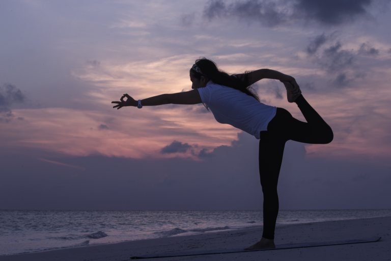 Silhouette einer Person in Yogapose am sandigen Strand mit Meer und rosa-violettem Himmel im Hintergrund