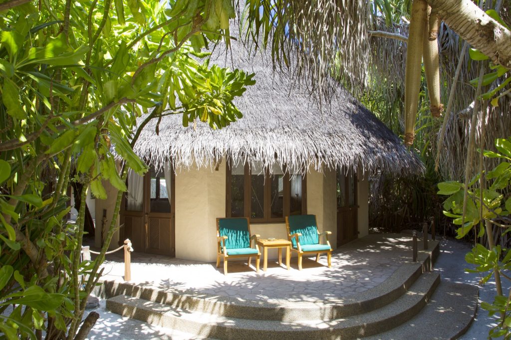 Eingangsbereich einer Beach Villa mit Strohdach, zwei grünen Holzliegestühlen und kleinem Holztisch