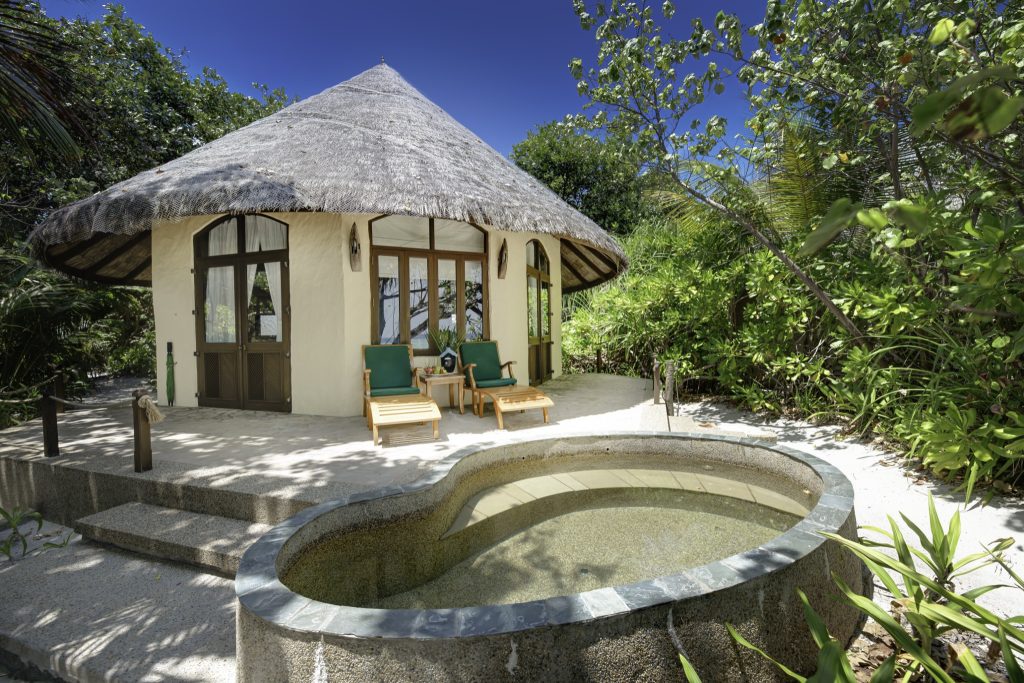 Runde Beach Villa mit Strohdach, Holzliegen mit grünen Polstern, kleinem Pool und üppigem Grün auf den