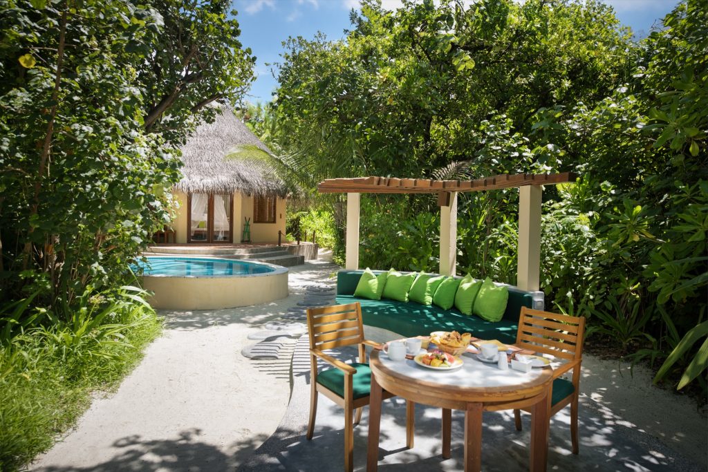 Deluxe Pool Villa mit Strohdach-Hütte, Holzsitzgruppe und Frühstück unter Holzpergola, umgeben von grüner Vegetation