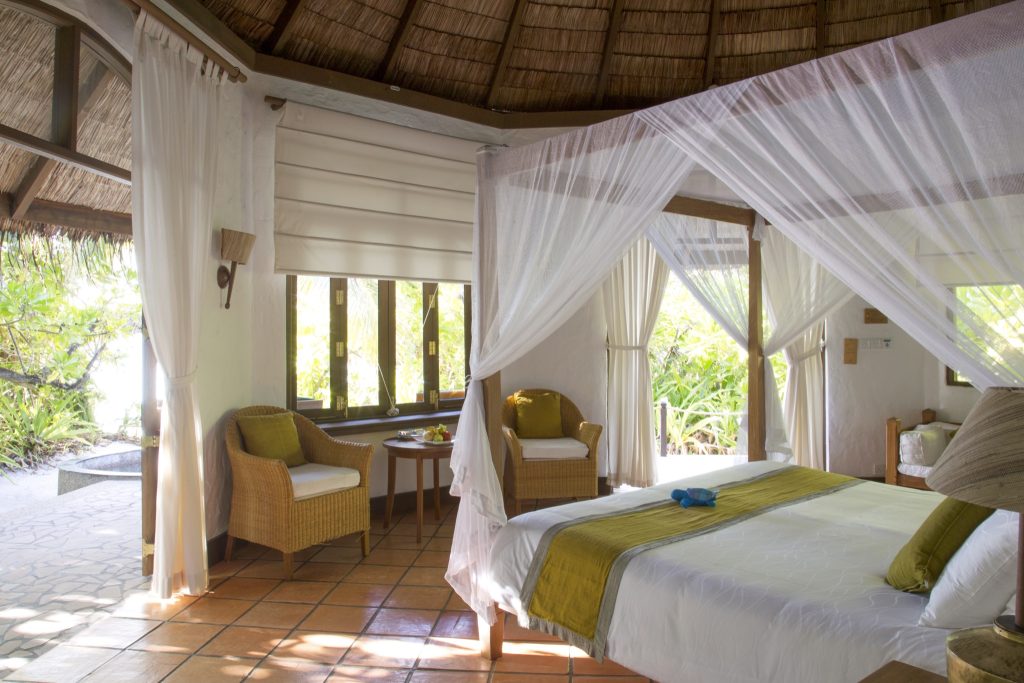 Zimmer der deluxe villa mit Himmelbett, Moskitonetz, Korbsesseln, Holztisch und großen Fenstern mit Blick auf Grün