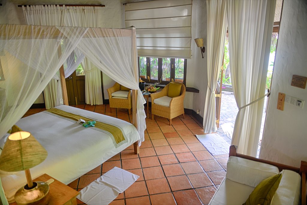 Helles Deluxe Villa Zimmer mit Himmelbett, Terrakottafliesen, Rattan-Sesseln und Terrasse in der Coco Palm Dhuni Kolhu