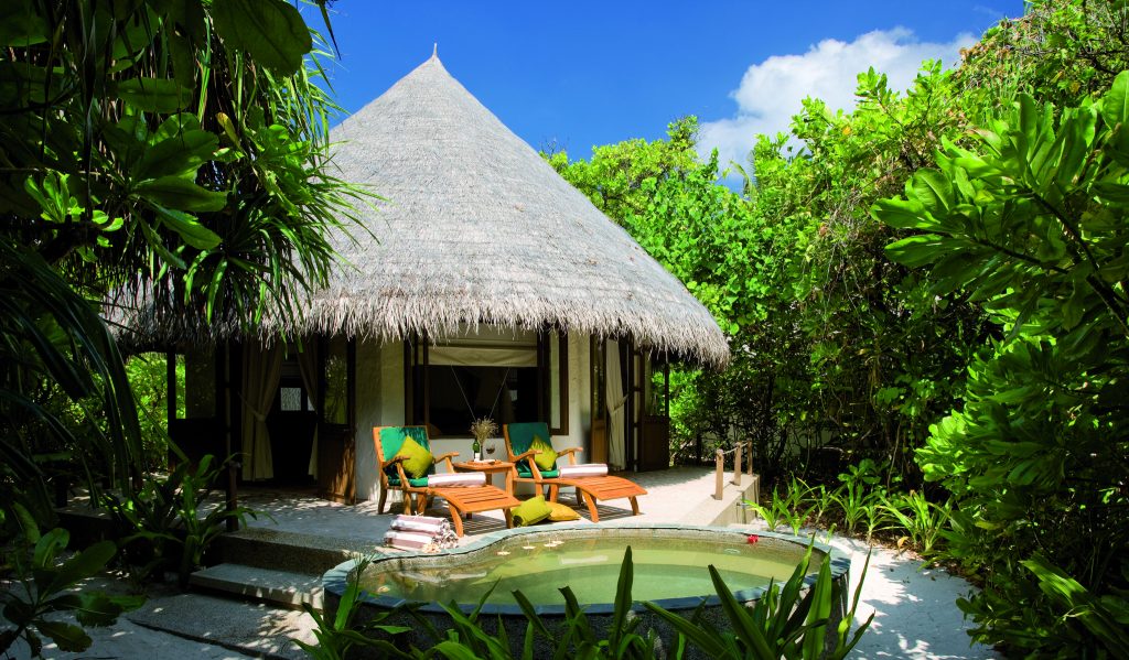 Deluxe Villa mit Reetdach, zwei hölzerne Liegestühle mit grünen Polstern vor kleinem Pool und grüner Vegetation