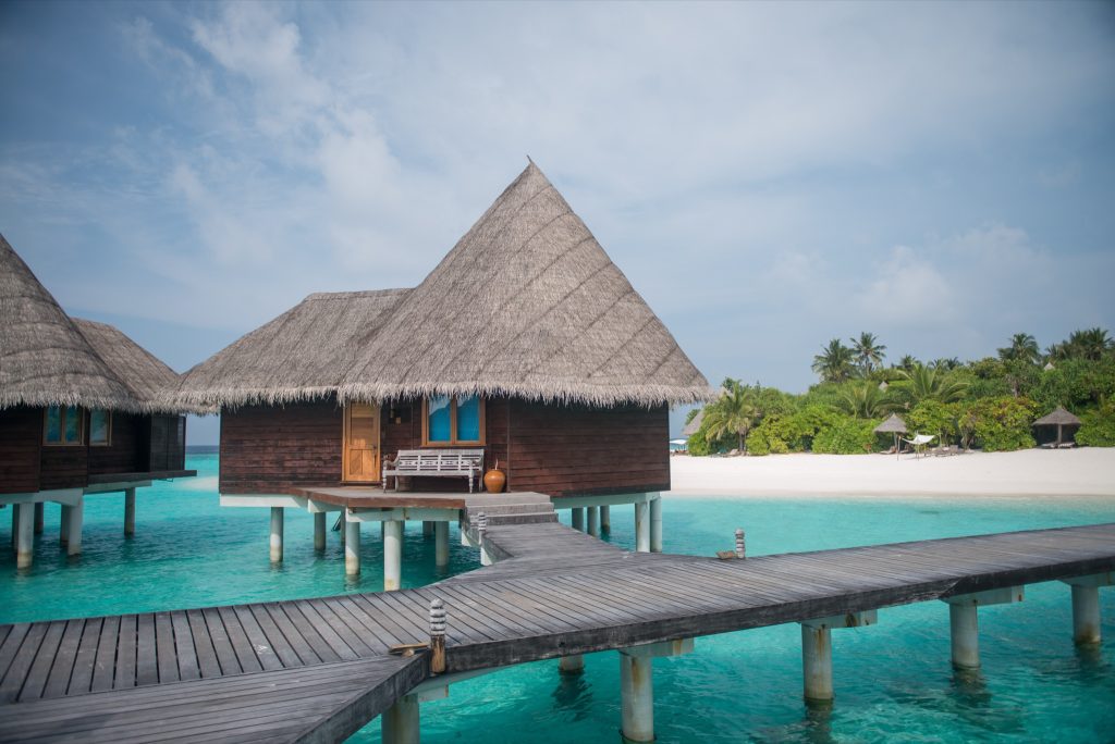 Wasserbungalows aus dunklem Holz mit Palmdächern auf Stelzen über türkisblauem Wasser, verbunden durch