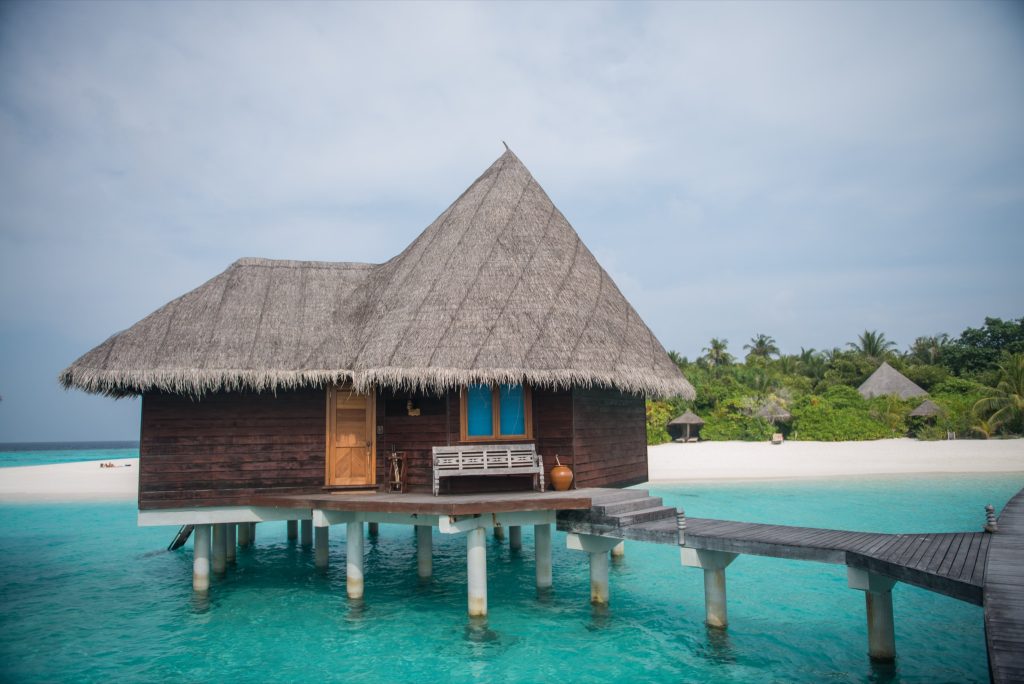 Lagoon Villa auf Stelzen mit dunklem Holz und Strohdach, Veranda mit Bank, Steg und türkisfarbenem Wasser auf den