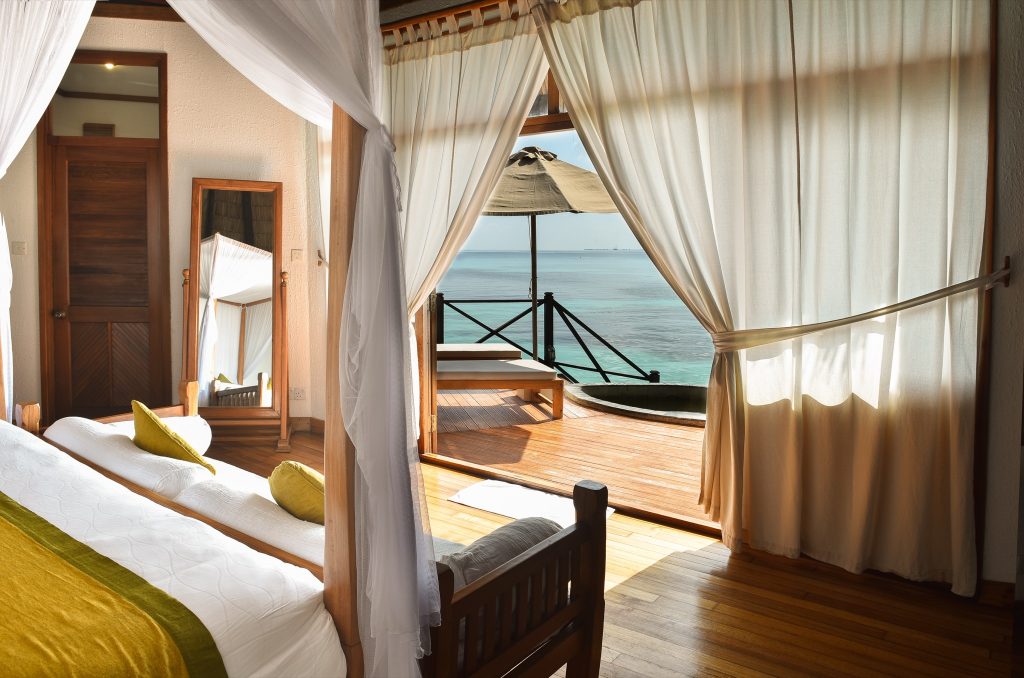 Zimmer der Lagoon Villa mit Himmelbett, weißen Vorhängen, Holzboden und Meerblick auf den Balkon