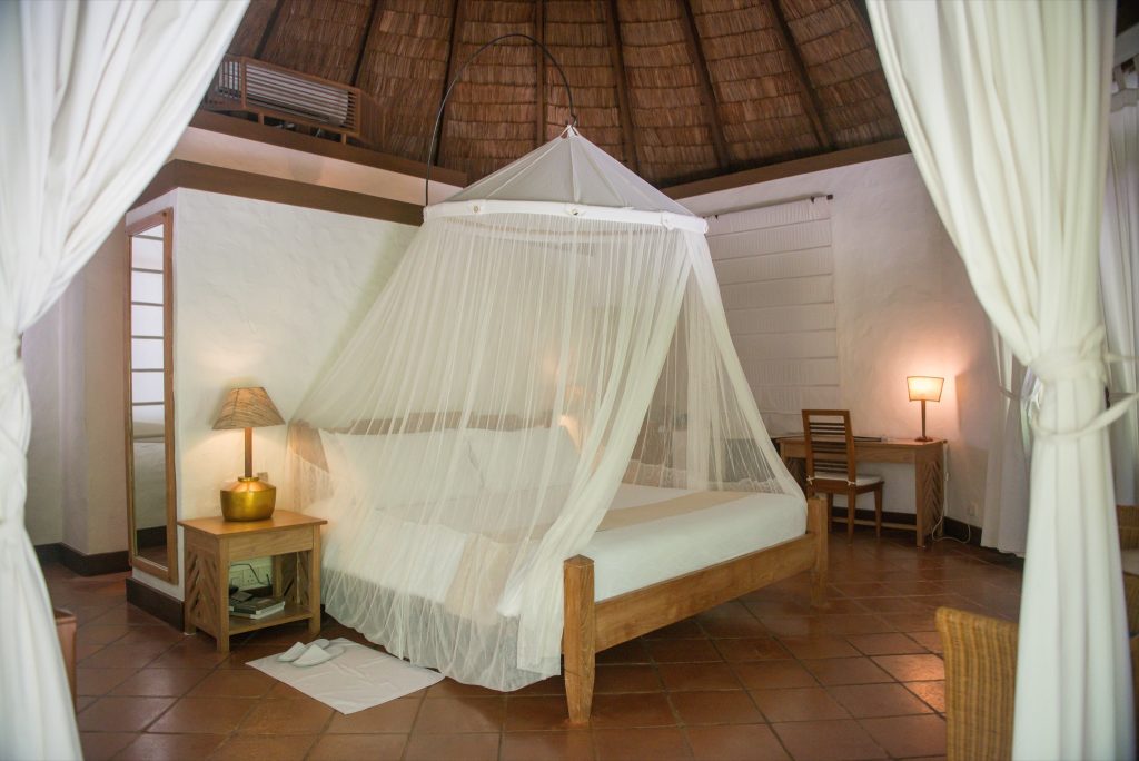 Geräumiges Zimmer der Sunset Beach Villa mit großem Bett und weißem Moskitonetz, Holzmöbeln und Strohdach