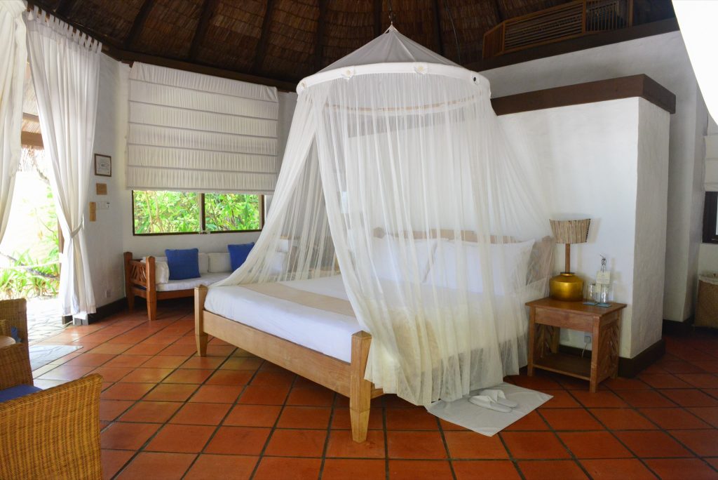 Zimmer der Sunset Beach Villa mit Himmelbett, Moskitonetz, Holzdecke, Terrakottafliesen und Sofa mit blauen Kissen