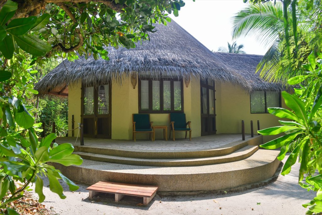 Bungalow der Sunset Beach Villa mit reetgedecktem Dach, gelben Wänden und grüner Veranda mit Holzmöbeln in tropischer.