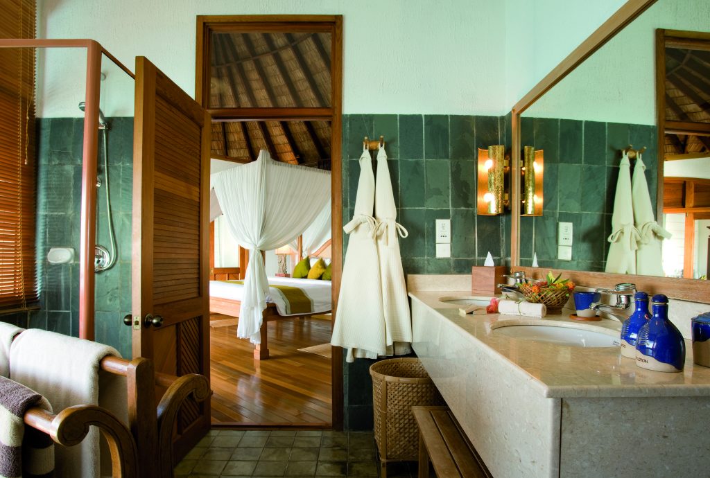 Badezimmer mit grünen Kacheln und Waschtisch aus hellem Stein, angrenzend an Schlafzimmer mit weißem Baldachin im Coco Palm.