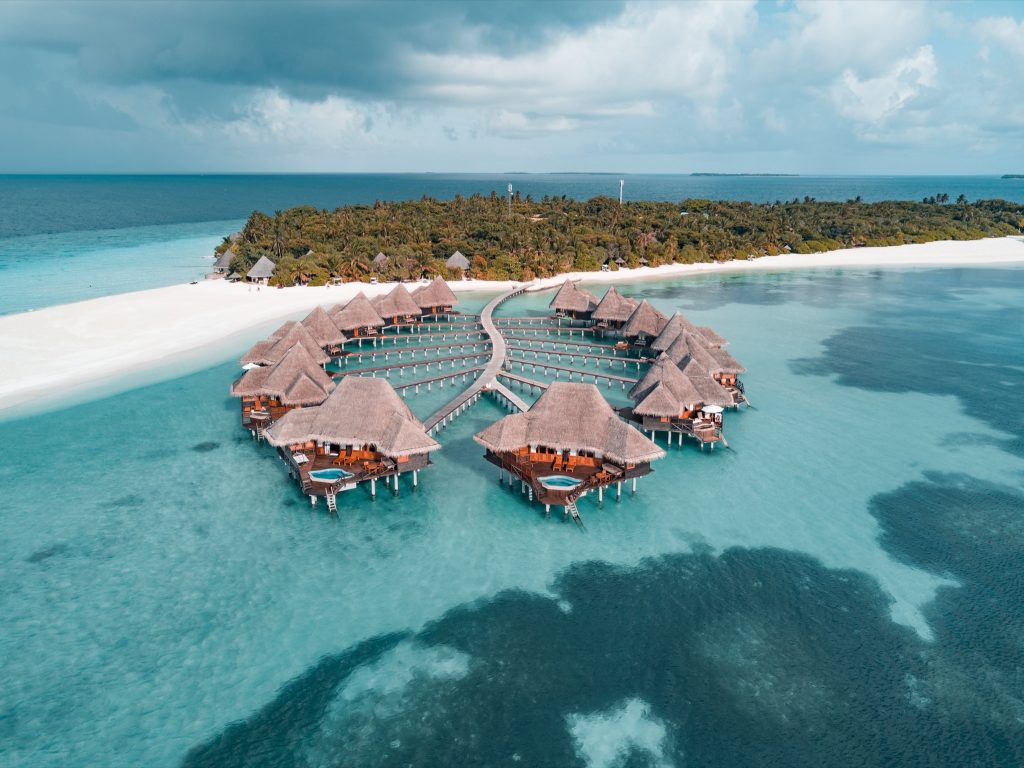 Wasserbungalows mit strohgedeckten Dächern auf Pfählen über türkisfarbenem Meer mit Stegen und weißem Sandstrand.