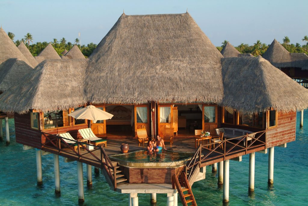 Bungalow auf Stelzen über türkisblauem Wasser mit Holzterrasse, Liegestühlen und Sonnenschirm auf den
