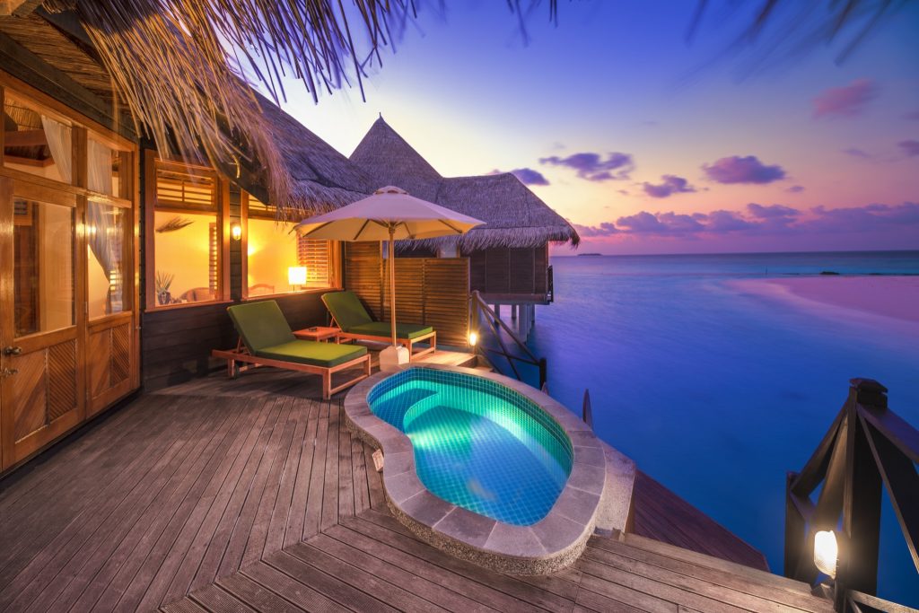 Terrasse eines Wasserbungalows mit beleuchtetem Pool, Holzplanken, grünen Liegestühlen und Meer bei Sonnenuntergang