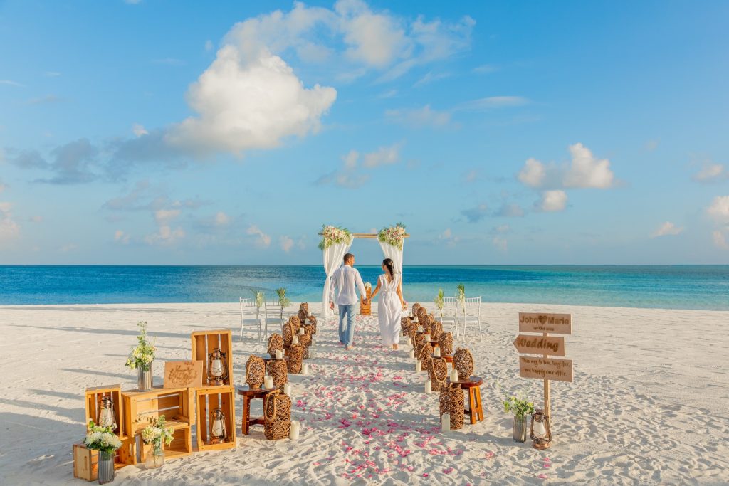 Hochzeitsszene am Strand mit Paar auf Sandweg, Holzbogen, weißen Dekorationen und Meereshorizont