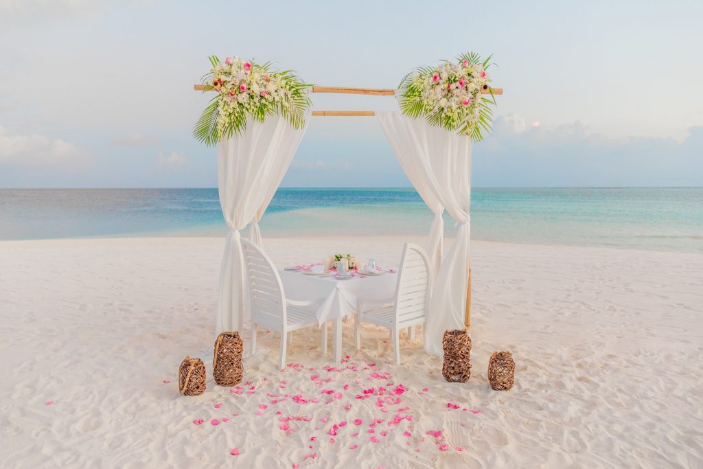 Holzbaldachin mit weißen Vorhängen und pinken Blumen an einem Strand mit Tisch