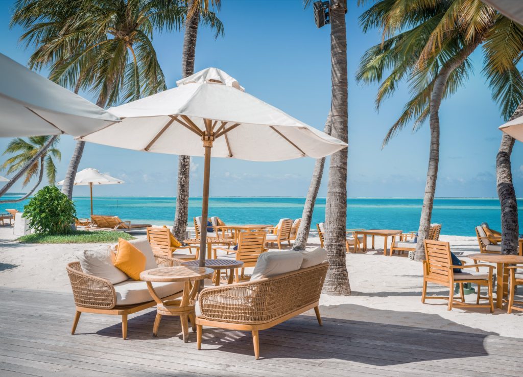 Holz- und Rattan-Sitzgarnituren mit weißen Kissen unter großen weißen Sonnenschirmen am Meer im Conrad Maldives Rangali Bar