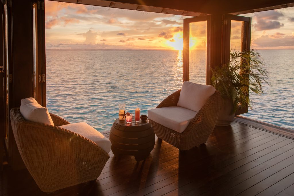 Gemütliche Bar-Sitzecke mit runden Rattansesseln und Holztisch am Meer bei Sonnenuntergang auf den