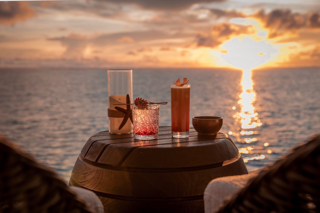 Runder Holztisch auf Terrasse mit zwei dekorierten Gläsern, Kerze mit Seestern und Blick auf Meer bei Sonnenuntergang