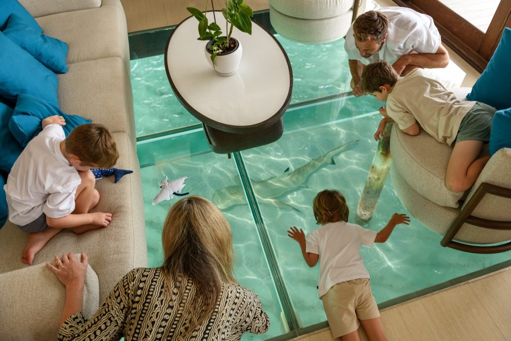Raum im Ocean Pavilion mit Glasboden, durch den ein Hai im Wasser schwimmt, umgeben von beigen Sofas mit blauen Kissen