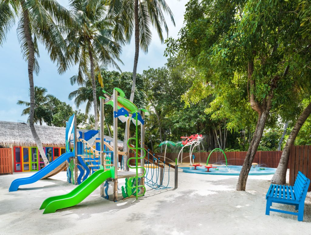 Bunter Spielplatz mit Klettergerüsten, Rutschen und Wassersprinklern umgeben von Palmen und tropischer Vegetation