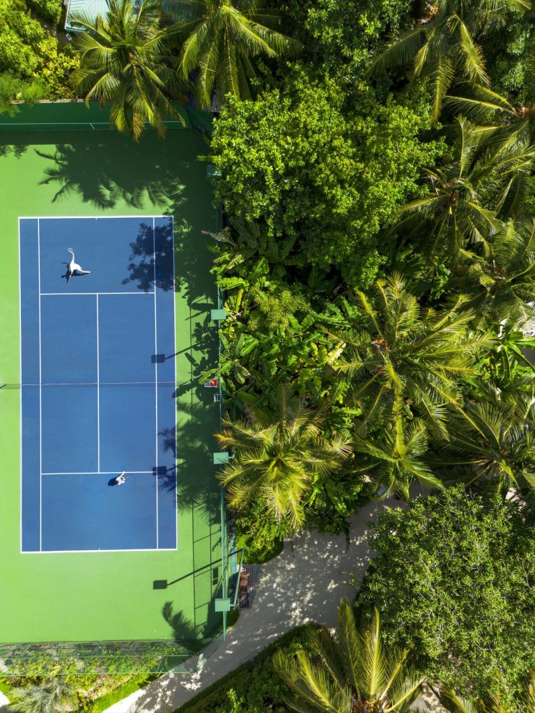 Tennisplatz mit blauem Kunststoffbelag und grünen Linien umgeben von tropischer Vegetation auf den